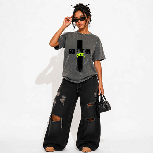 A woman modeling a washed charcoal gray graphic t-shirt with distressed black cross that reads ‘God is within’ in black ‘HER’ in neon green, wide-leg jeans, sunglasses, and a small black handbag against a white studio background.