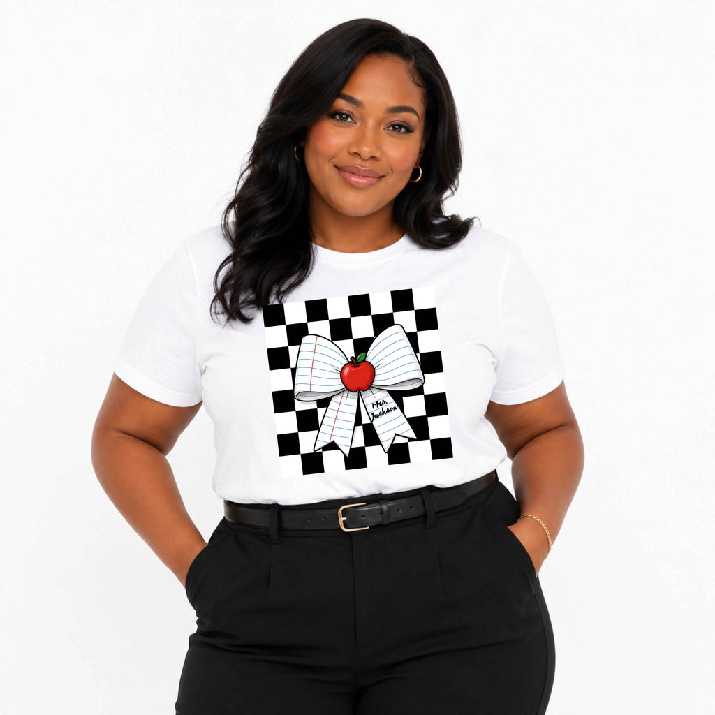 A woman modeling a white short-sleeve t-shirt with a black and white checkerboard graphic background and a large white bow made of notebook paper with a red apple in the center. She is wearing black pants and a black belt against a plain white studio background.