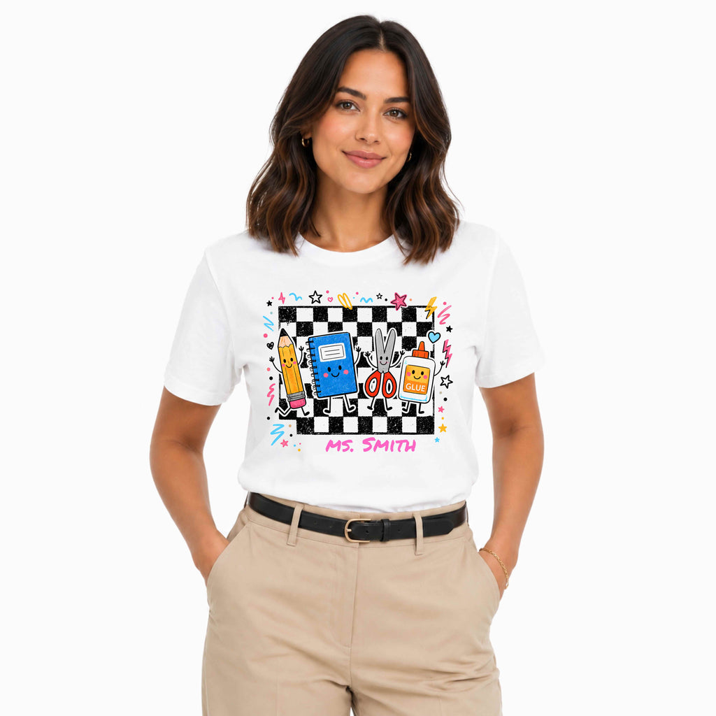 A woman modeling a white short-sleeve graphic T-shirt with a colorful cartoon-style chest print featuring a black-and-white checkerboard, a blue notebook, dancing cartoon supplies, and the text 'MS. SMITH', paired with beige pants against a light gray studio background.