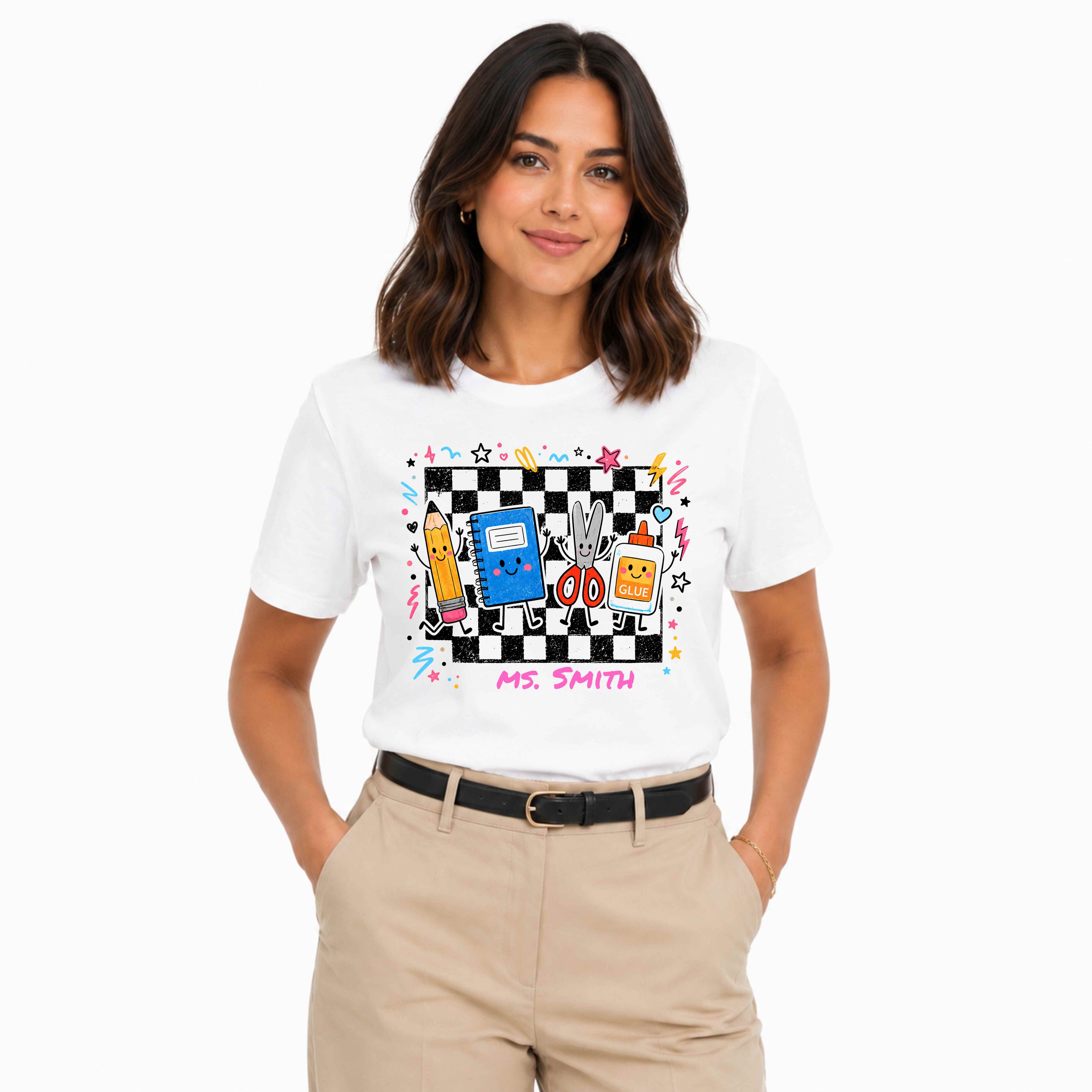 A woman modeling a white short-sleeve graphic T-shirt with a colorful cartoon-style chest print featuring a black-and-white checkerboard, a blue notebook, dancing cartoon supplies, and the text 'MS. SMITH', paired with beige pants against a light gray studio background.