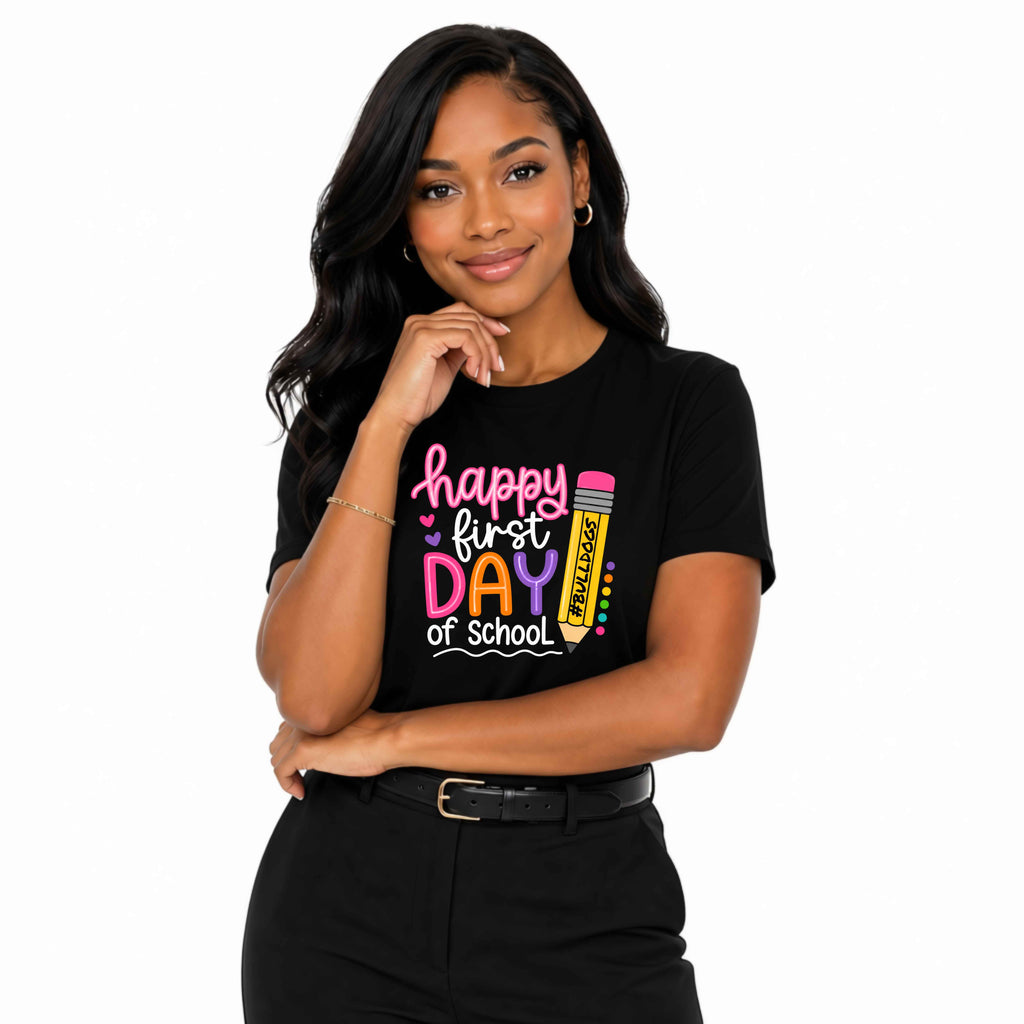 A woman modeling a black short-sleeve t-shirt with a colorful graphic that reads 'happy first DAY of school' alongside a pink and yellow pencil illustration, set against a plain white background.