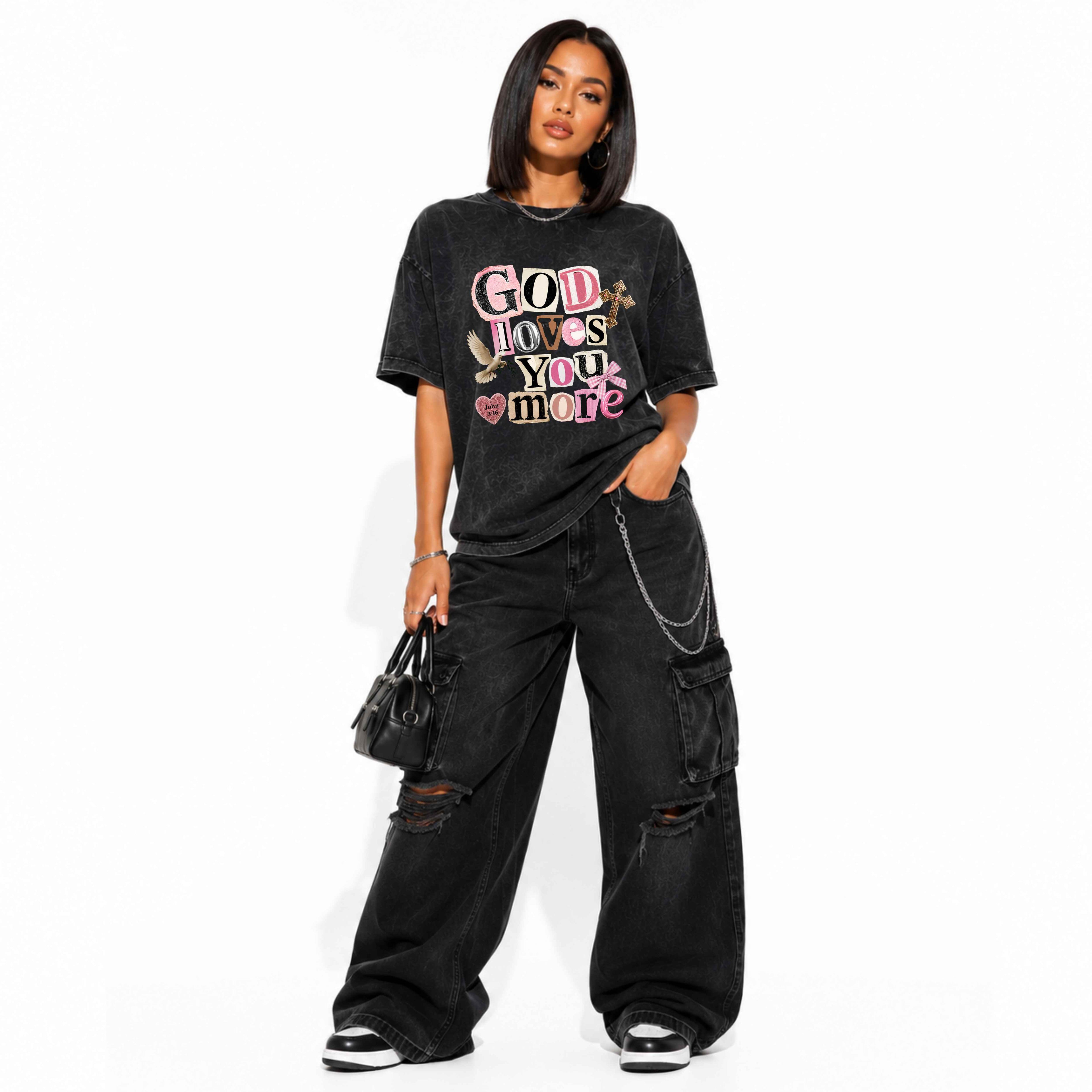 A woman modeling a black acid-wash graphic t-shirt with the text 'God Loves You More' in pink, white, and yellow, paired with black wide-leg cargo pants and holding a small black handbag against a white background.