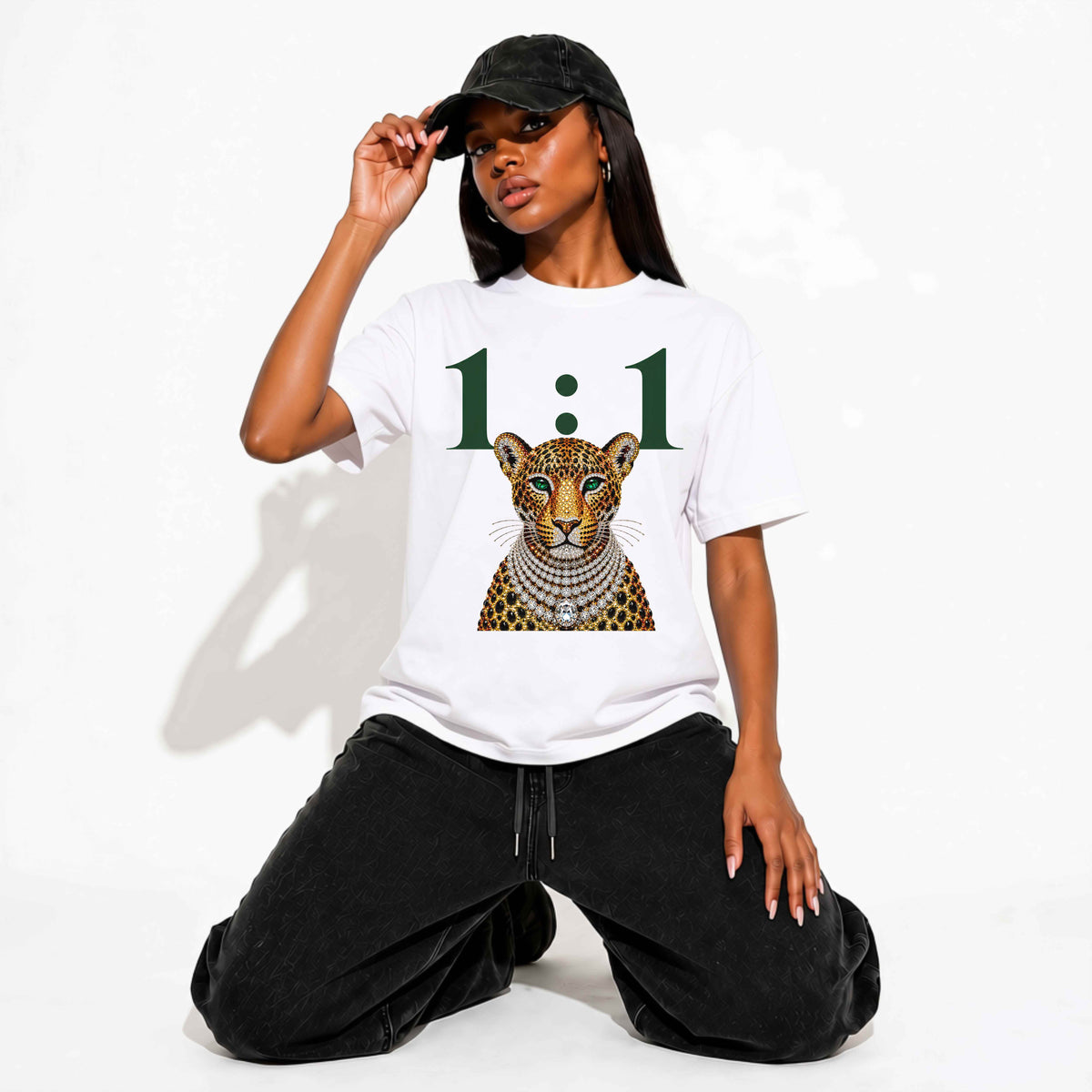 A woman kneeling on a white studio background wearing a white graphic T-shirt with a large green '1:1' and a jeweled leopard print, paired with black pants and a black cap.