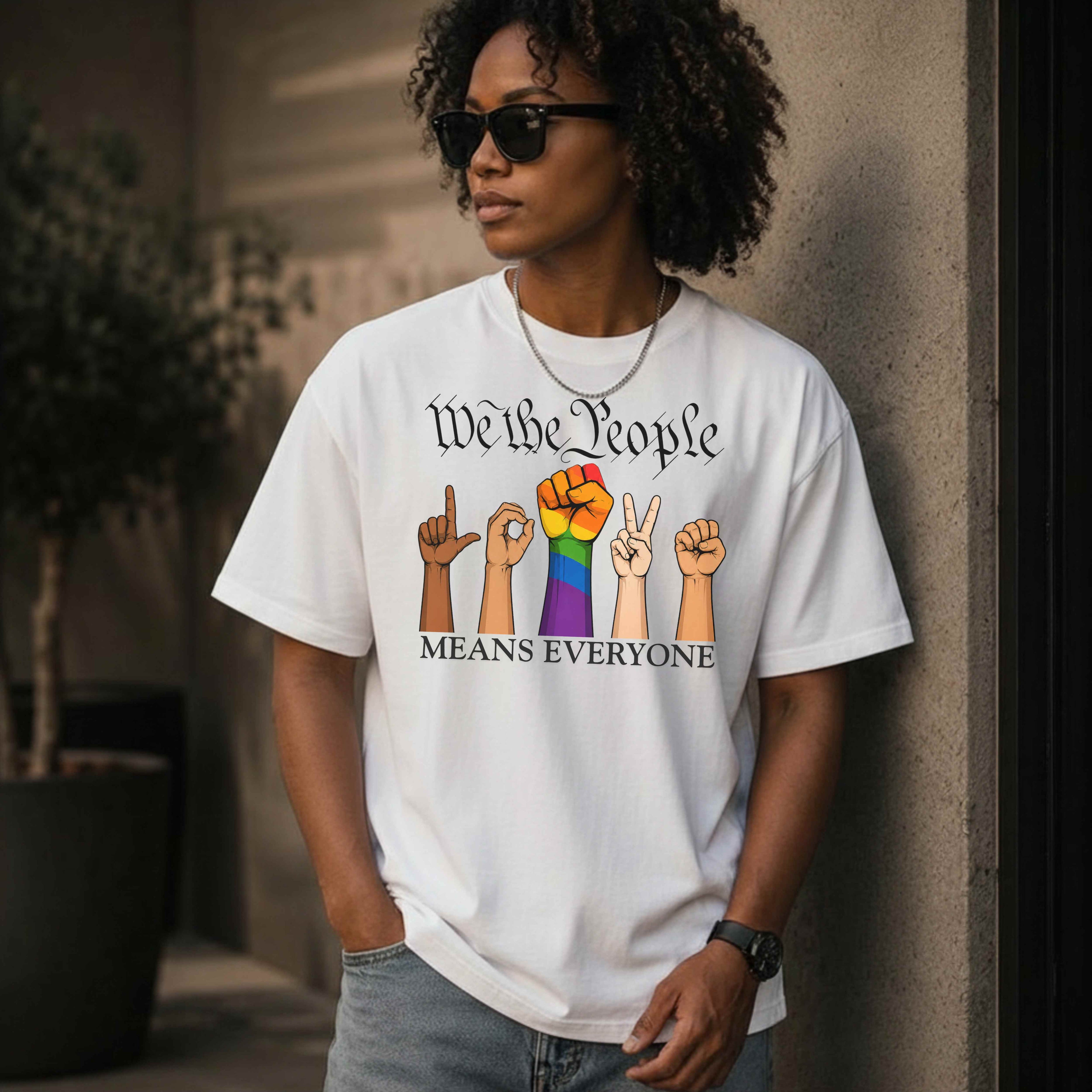 White mockup unisex graphic T-shirt featuring the phrase "We The People Means Everyone" with diverse illustrated hands spelling "LOVE" in sign language, an LGBTQ+ rainbow  raised fist in between. Inclusive Pride and equality statement shirt for everyday wear.
