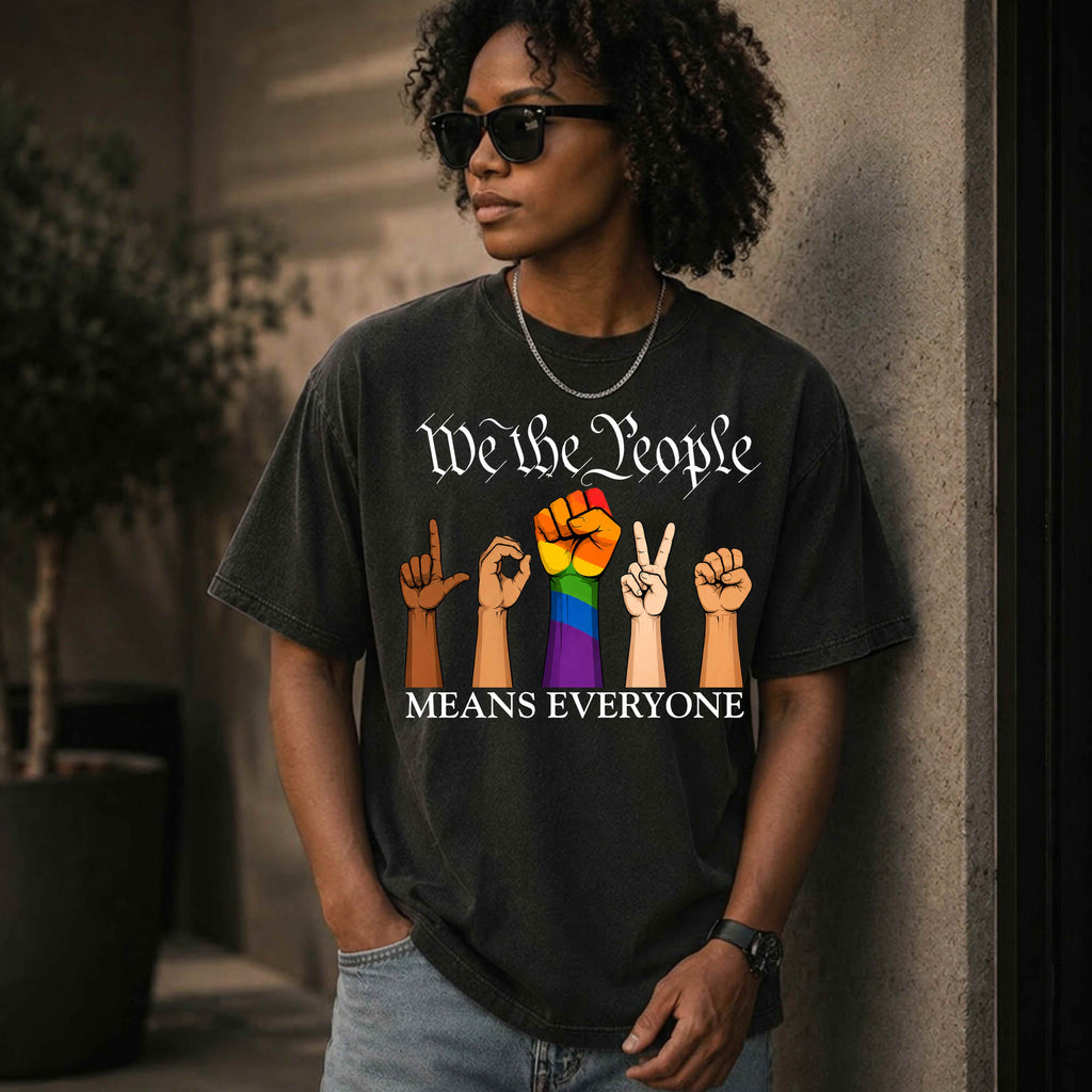 Black unisex graphic T-shirt featuring the phrase "We The People Means Everyone" with diverse illustrated hands spelling "LOVE" in sign language, an LGBTQ+ rainbow  raised fist in between. Inclusive Pride and equality statement shirt for everyday wear.