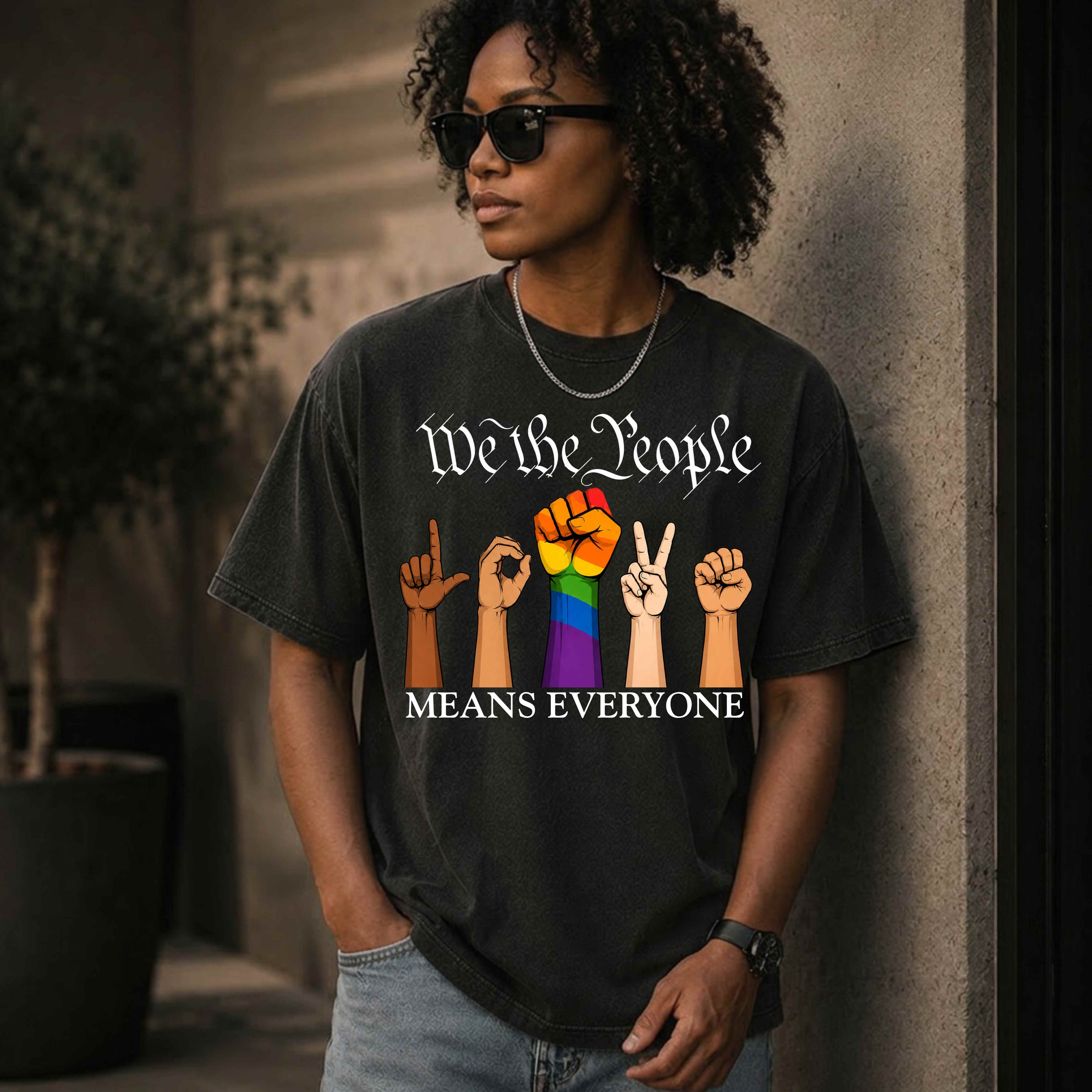 Black unisex graphic T-shirt featuring the phrase "We The People Means Everyone" with diverse illustrated hands spelling "LOVE" in sign language, an LGBTQ+ rainbow  raised fist in between. Inclusive Pride and equality statement shirt for everyday wear.