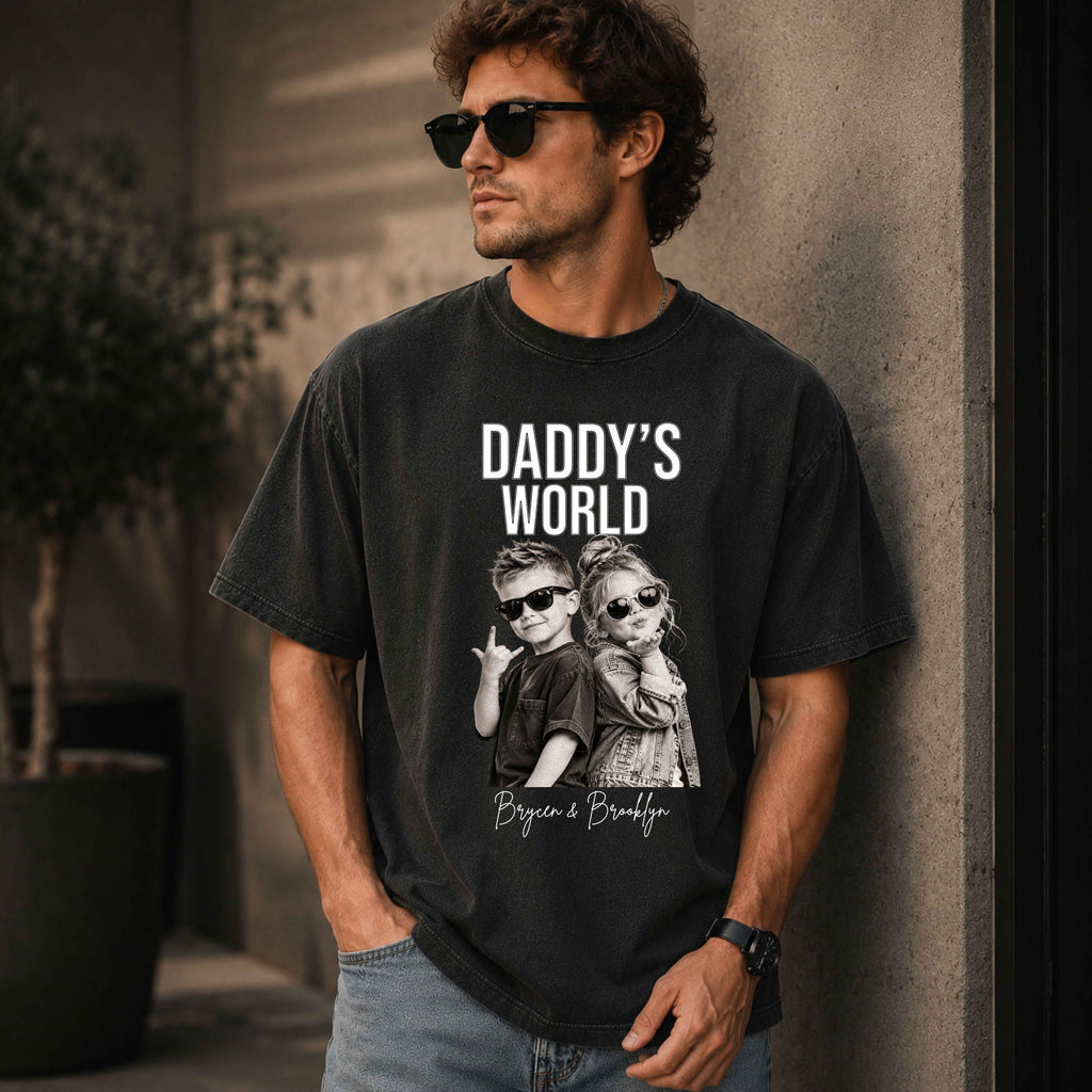 Man wearing a black t-shirt with 'Daddy's World' graphic with pictures of 2 children for personalization, standing against a wall.